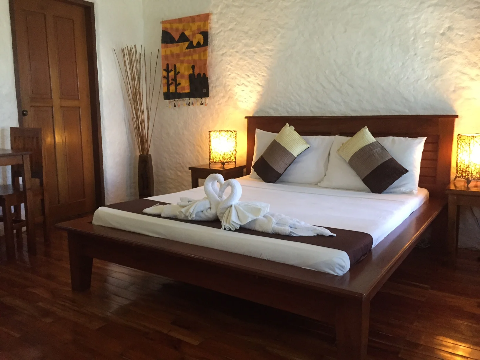 Cozy hotel room with wooden bed, white linens, decorative pillows, and towel art on bed, warm lighting from lamps.